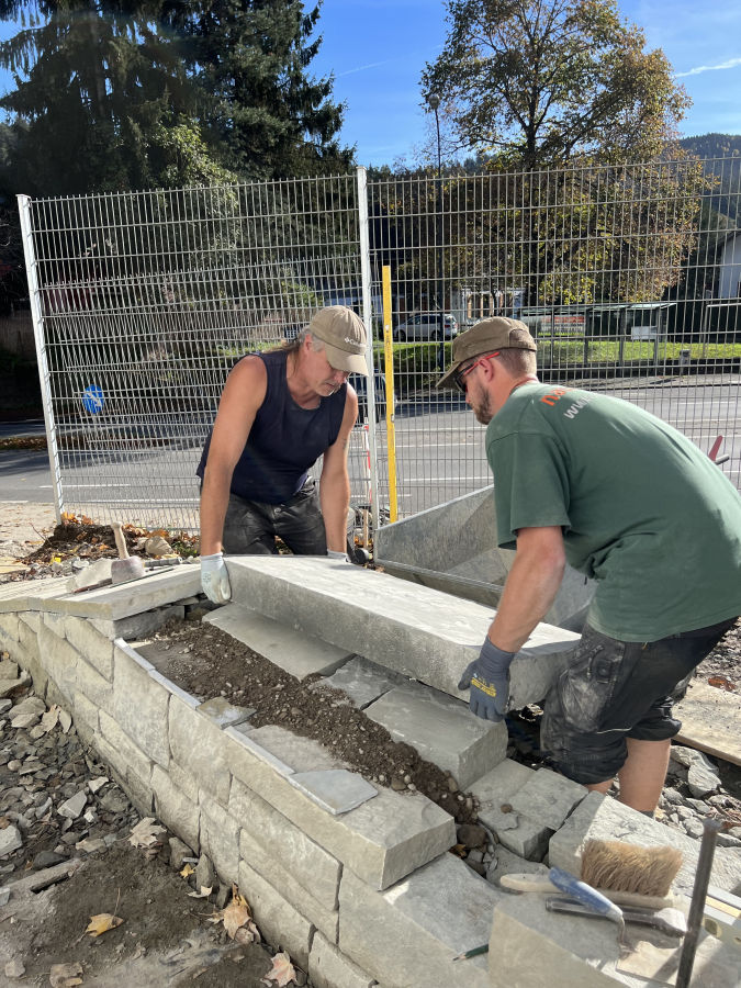 Ulf (Steinarbeiter) und Max (Naturgärtner) bauen einen schweren Mauerdeckel ein.